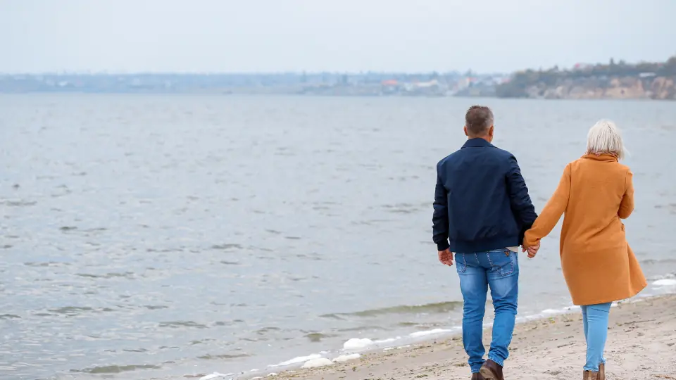 Partnersuche über Dating Seiten ab 50: ein älteres Paar geht Hand in Hand am Strand entlang
